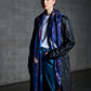 A full-length photo of a woman posing in an industrial-style setting, wearing a long black leather coat, a white t-shirt, and blue jeans. Her look is accessorized with a vibrant dark blue scarf featuring colorful space-themed illustrations, which hangs freely in front. The minimalist lighting and the concrete wall in the background create a modern and edgy aesthetic, highlighting the stylish outfit.
