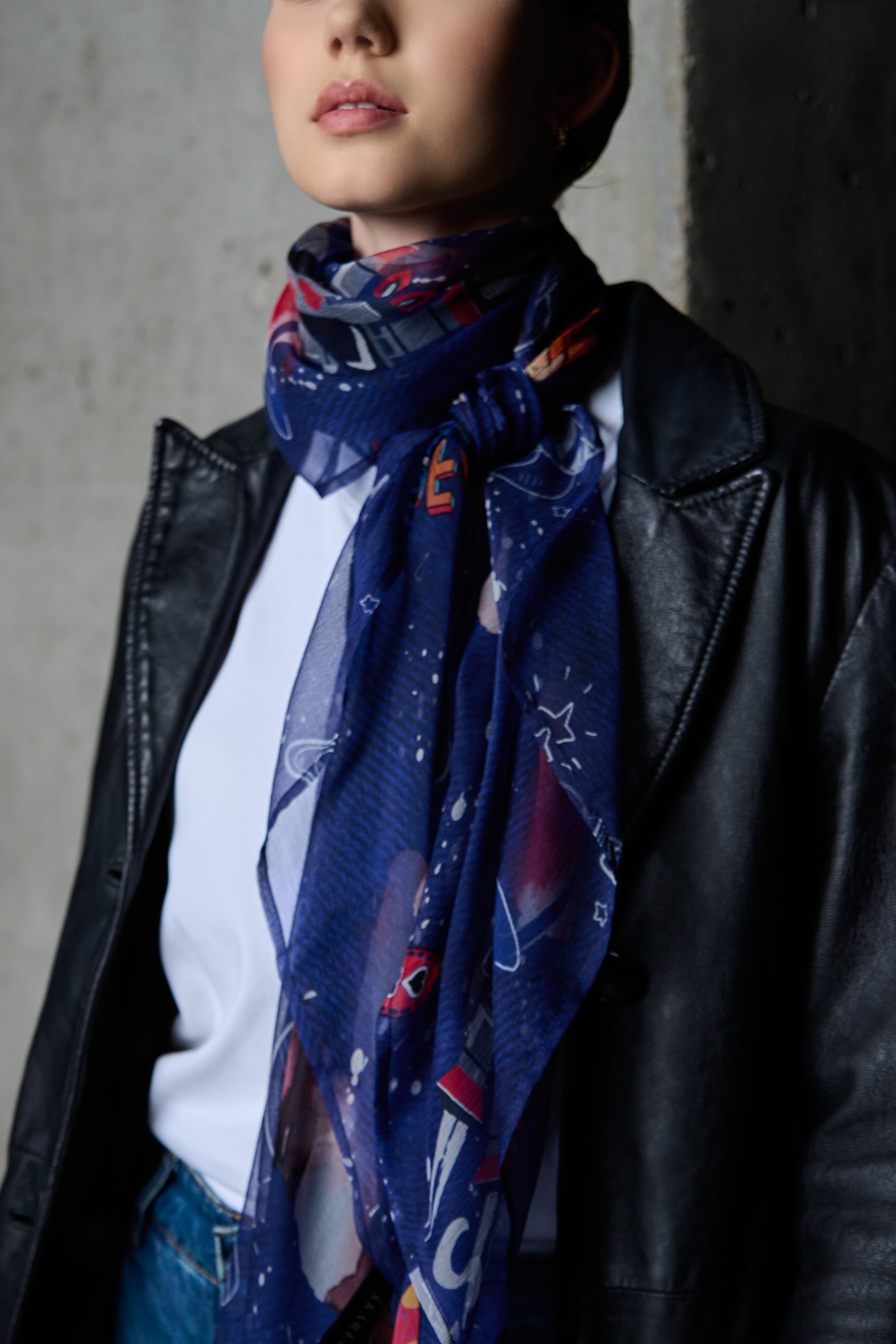 A close-up photo showing a woman's chest and neck adorned with a dark blue silk scarf featuring colorful space-themed illustrations. The scarf is stylishly tied in a knot, with its loose ends falling over a white t-shirt. The woman is wearing a black leather jacket, and a grey concrete wall is visible in the background. The focus is on the details of the accessory and the texture of the fabric, creating a modern and elegant aesthetic.