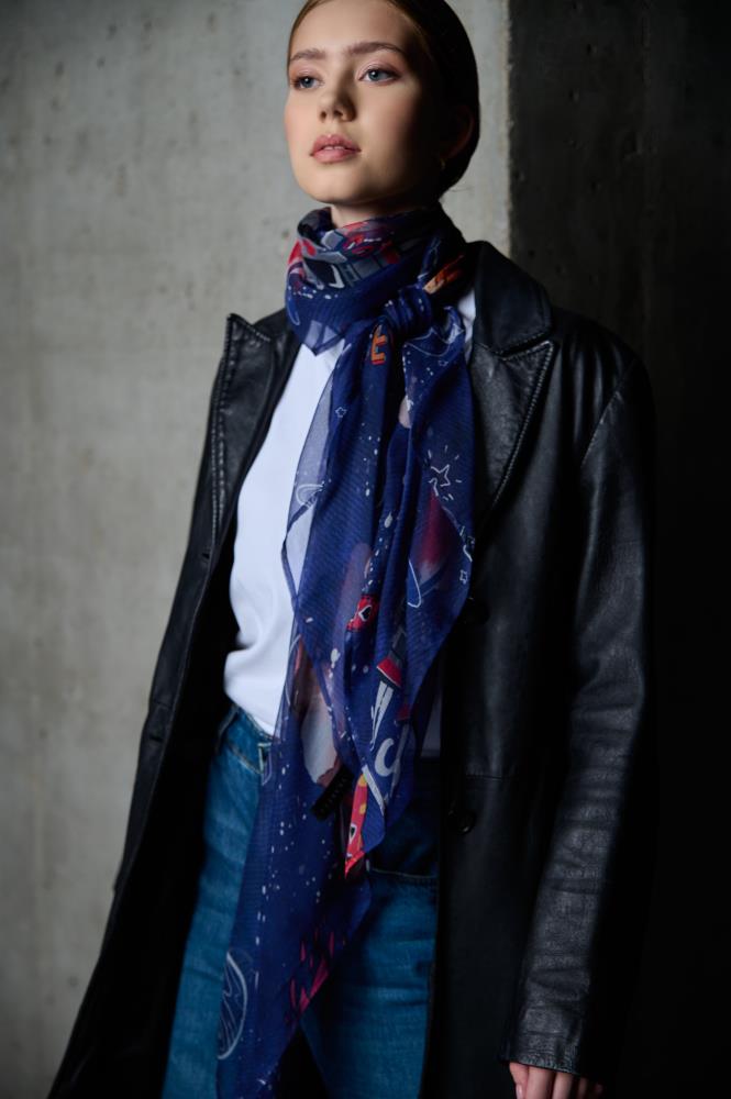 A medium shot of a woman posing against a concrete wall in an industrial setting. She is wearing a black leather coat over a white t-shirt and blue jeans. A dark blue scarf, decorated with colorful space-themed motifs like planets, rockets, and stars, is stylishly tied around her neck. The lighting highlights the model's face and the contrasting textures of the fabrics – the matte leather and the lightweight material of the scarf.