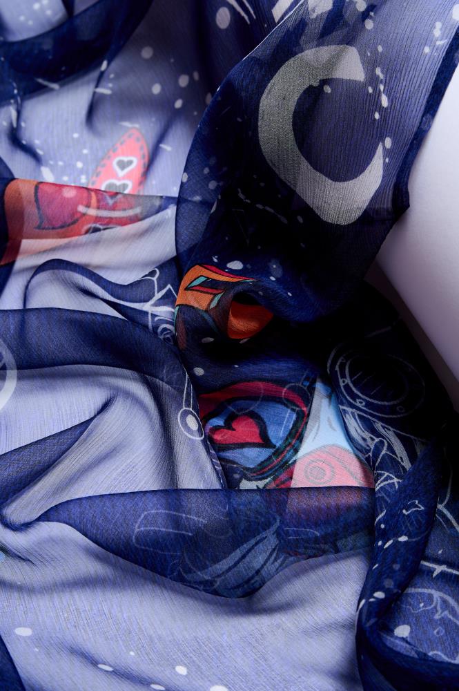 An extreme close-up showing the folds of a dark blue, sheer scarf. The fabric is decorated with playful and colorful space-themed illustrations: a grey crescent moon, a red rocket with hearts, and other abstract drawings are visible. The photo highlights the light, delicate texture of the fabric, its transparency, and how the material plays with light, creating a sense of depth and softness.