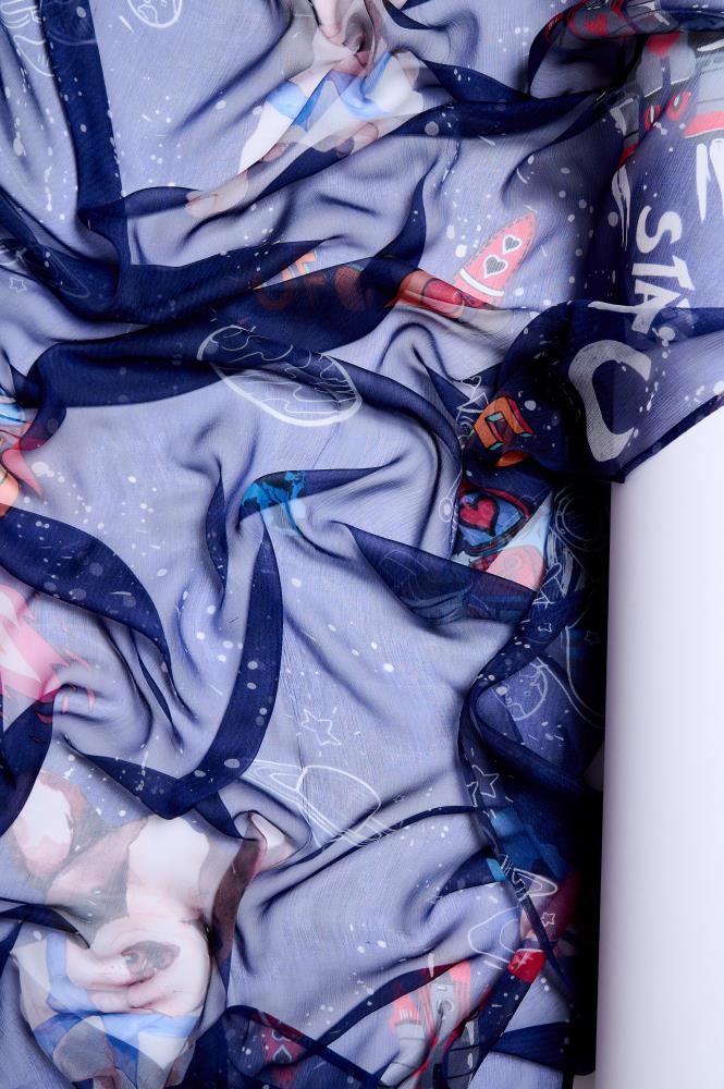 A close-up photo showing the folds of a dark blue, lightweight, and sheer scarf artistically laid out on a white background. The fabric is decorated with colorful and playful space-themed illustrations: planets with rings, rockets, stars, and cartoon-style characters. Soft lighting highlights the delicate texture of the fabric, its transparency, and the vibrant colors of the pattern, creating a cozy and creative aesthetic.