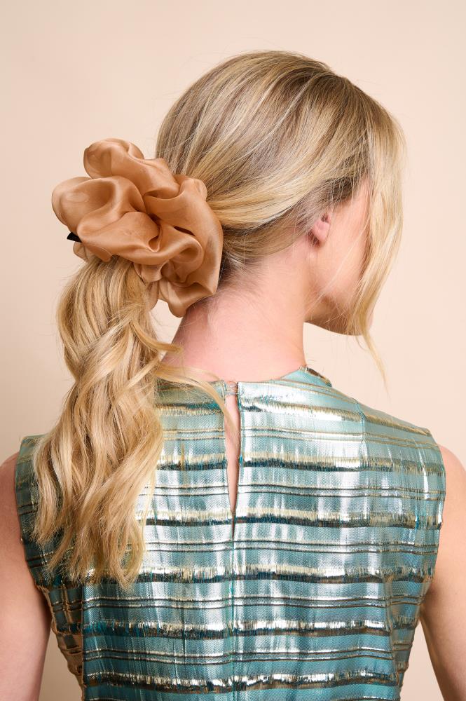A back view of a woman with blonde wavy hair tied in a ponytail. Her hair is styled with an oversized, voluminous golden-beige sheer organza silk scrunchie. She is wearing a shimmering blue and gold striped top against a light, neutral background.