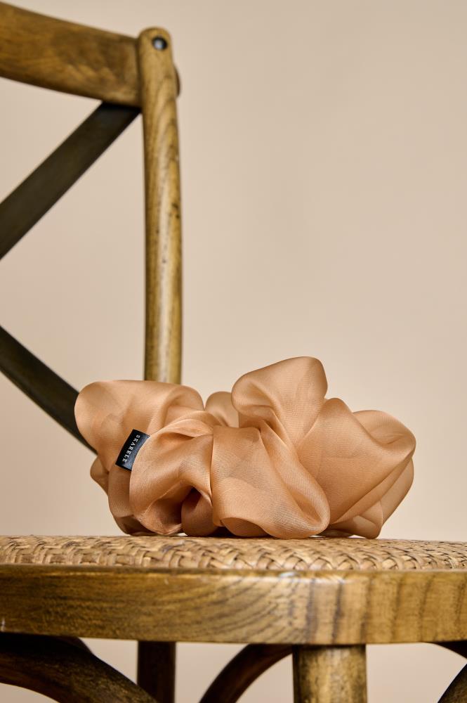 A voluminous, peach color silk organza scrunchie placed on the woven seat of a wooden chair. The light and abundant fabric folds give the accessory an elegant, airy look. The background is warm and neutral, and the natural wood texture creates a sense of coziness.
