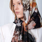 Woman wearing a floral patterned headscarf and white blazer, gazing to the side. The scarf features earthy tones, creating an elegant and thoughtful look.