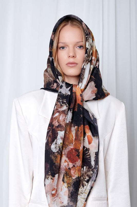Woman in a white blazer wearing a floral-patterned headscarf. She gazes forward with a neutral expression against a light-colored background.
