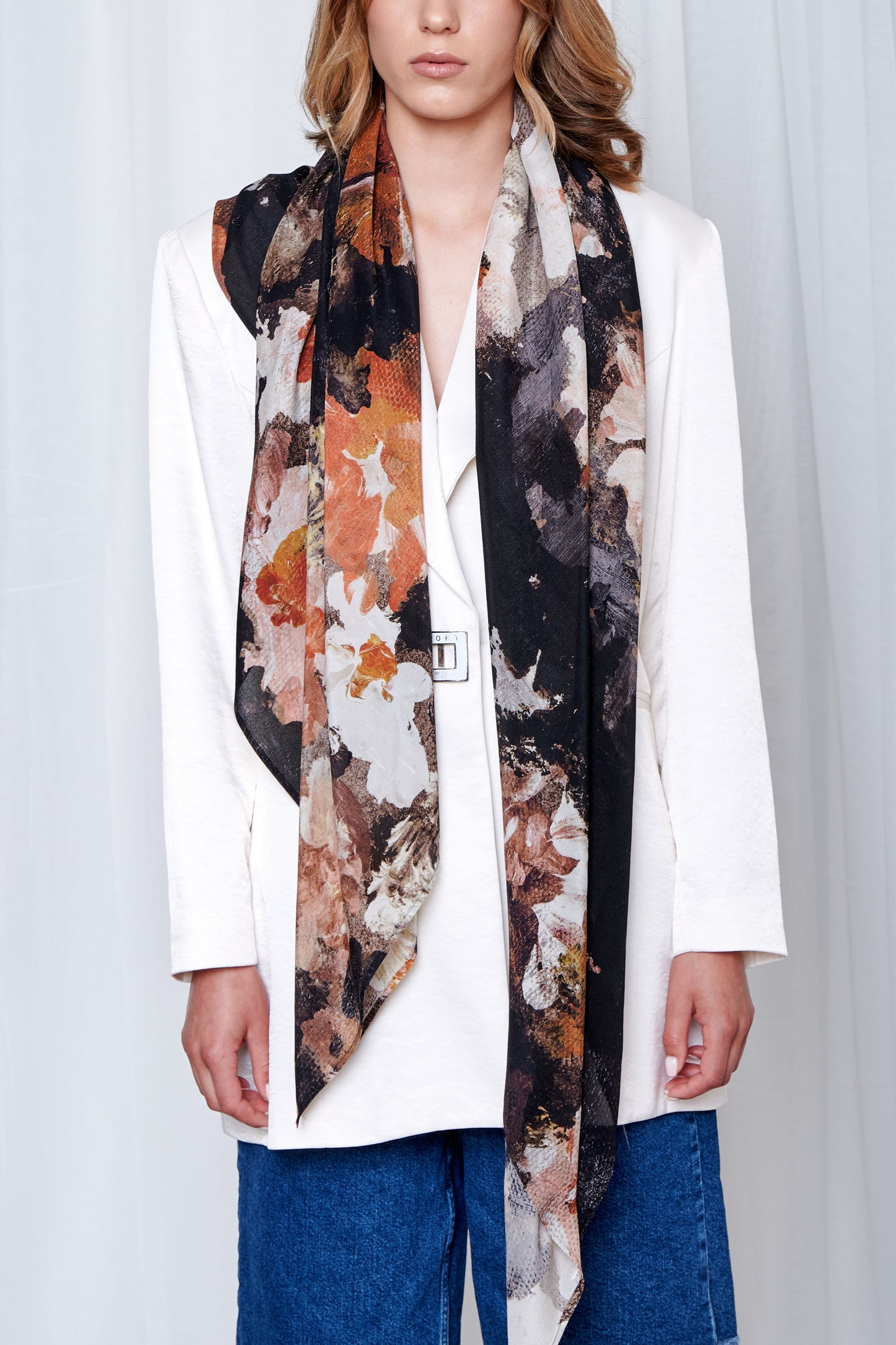 A person in a white blazer with a long, floral-patterned silk scarf in earthy tones, paired with blue jeans. The mood is stylish and elegant.