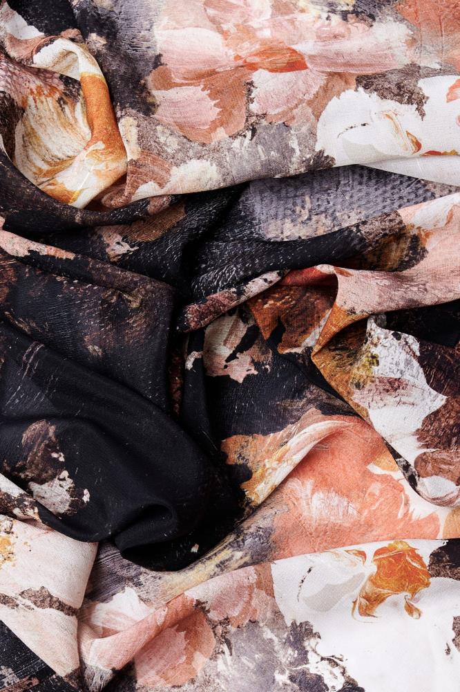 Close-up of crumpled silk fabric with an abstract pattern of earthy tones, including black, white, orange, and brown. The texture appears soft and rich.