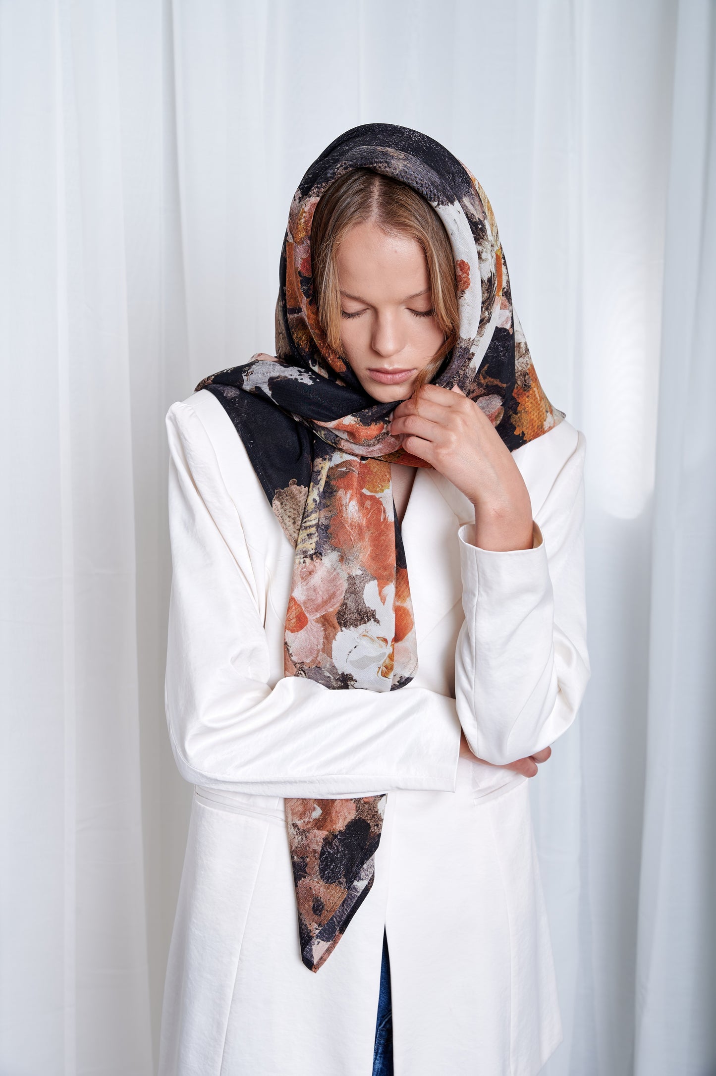 Woman in a white jacket and floral maxi silk scarf styled around her head like elegant hood stands against a white curtain, looking down with closed eyes. The tone is contemplative and serene.