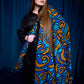 A young woman standing against dark blue curtains, wearing black clothing. She is wrapped in a large, vibrant blanket and wearing a matching headscarf. Both accessories feature a rich Baroque-style pattern in shades of royal blue, cyan, and bronze. Her long brown hair falls over her shoulders, accentuating the texture of the accessories.