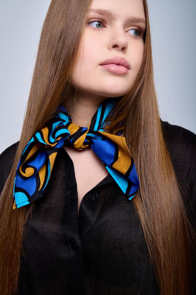 A portrait of a young woman with long, straight brown hair looking to the side. She is wearing a black shirt, accented by a vibrant cotton scarf tied around her neck. The scarf features a blue, cyan, and bronze Baroque-style pattern with bold black outlines.