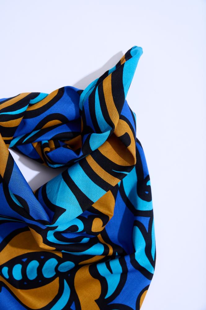 A close-up of a twisted and knotted fabric against a light background. The material features a dynamic Baroque-style pattern with royal blue, cyan, and bronze elements separated by bold black lines. The knot and folds create a playful display of shadows and texture.