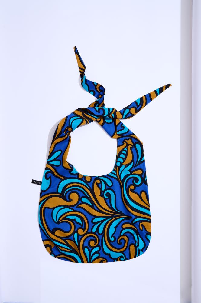A soft-shaped fabric tote bag photographed against a white background. The bag features a vibrant Baroque pattern with royal blue, cyan, and bronze motifs outlined in black. The handles are tied in a knot at the top, and a small black brand label is visible on the left side.