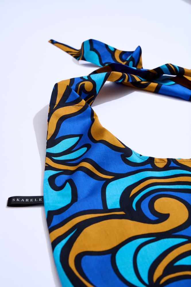 a close-up shot of a fabric tote bag against a white background. The bag features a vibrant pattern in shades of royal blue, cyan, and bronze with bold black outlines. A small black label with the white text "SKARELE" is visible on the left side.