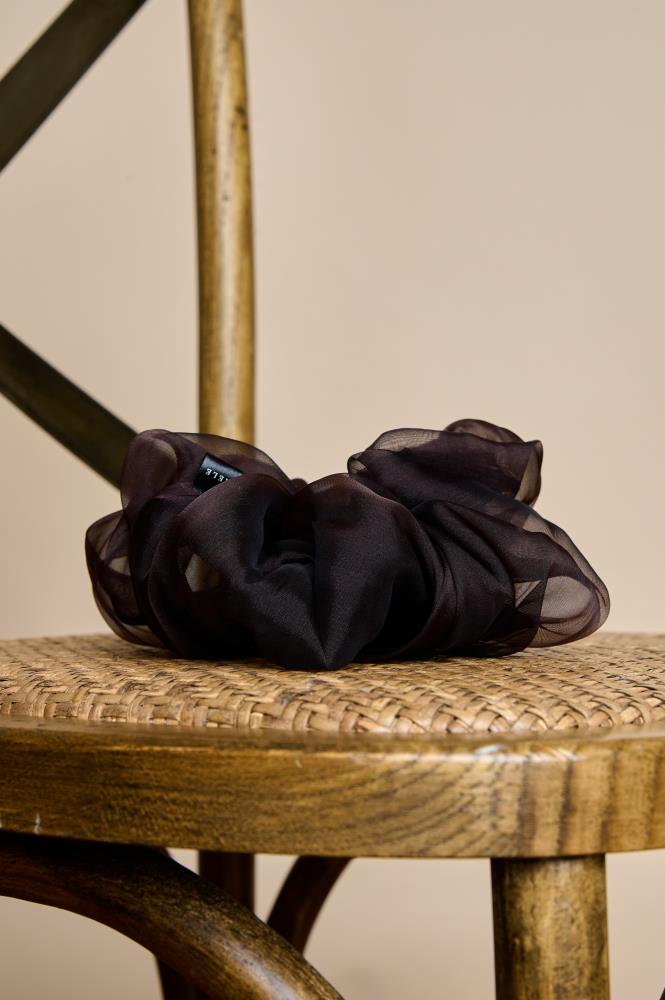 A voluminous, dark brown  sheer fsilk organza scrunchie placed on the woven seat of a wooden chair. The light folds of the fabric create an elegant, airy look, while the warm lighting and the chair's texture give the photo a cozy, rustic feel.