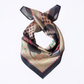 A square silk scarf with a complex abstract pattern featuring beige, brown, black, green, grey and red, tied in a knot against a plain white background.