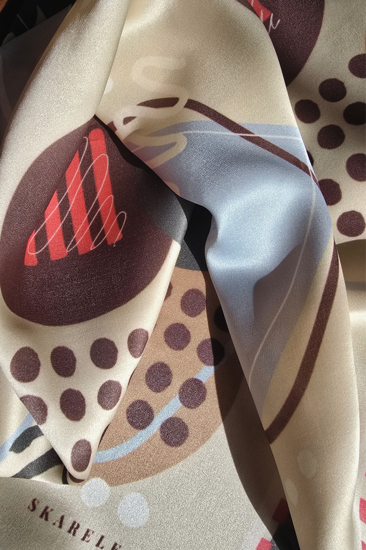 A close-up of a silk scarf with a modern abstract print featuring brown polka dots, a red striped motif, and a light blue wave on a beige background.