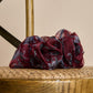 A voluminous, burgundy sheer fabric scrunchie placed on the woven seat of a wooden chair. The accessory is decorated with dark, animal-print motifs. The light fabric folds and warm lighting, combined with the natural wood texture, create a cozy, rustic atmosphere.