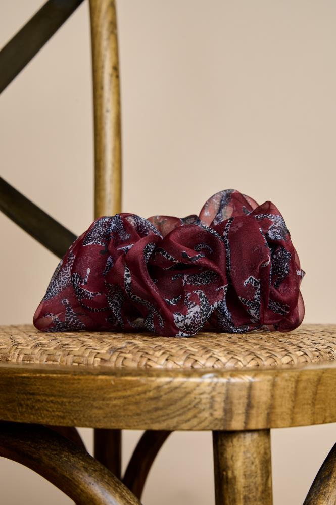 A voluminous, burgundy sheer fabric scrunchie placed on the woven seat of a wooden chair. The accessory is decorated with dark, animal-print motifs. The light fabric folds and warm lighting, combined with the natural wood texture, create a cozy, rustic atmosphere.