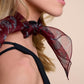 A close-up portrait of a woman showcasing a narrow burgundy silk scarf stylishly tied around her neck. The scarf features a subtle sheer texture and is decorated with a grey leopard pattern. An elegant accessory that adds luxury and classic charm to a minimalist look with a black top.