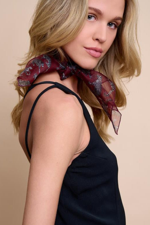 A side profile portrait of a model showcasing a narrow burgundy silk scarf with a leopard pattern. The scarf is stylishly tied around the neck, and its light, sheer fabric adds an elegant touch to the look. A black sleeveless top and a minimalist background highlight this luxurious accessory