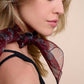 A profile photo of a model focusing on a stylish burgundy silk scarf with a leopard pattern. The scarf is tied around the neck, highlighting the light and sheer fabric texture. Minimalist makeup and soft lighting emphasize the elegant and luxurious image of this accessory.