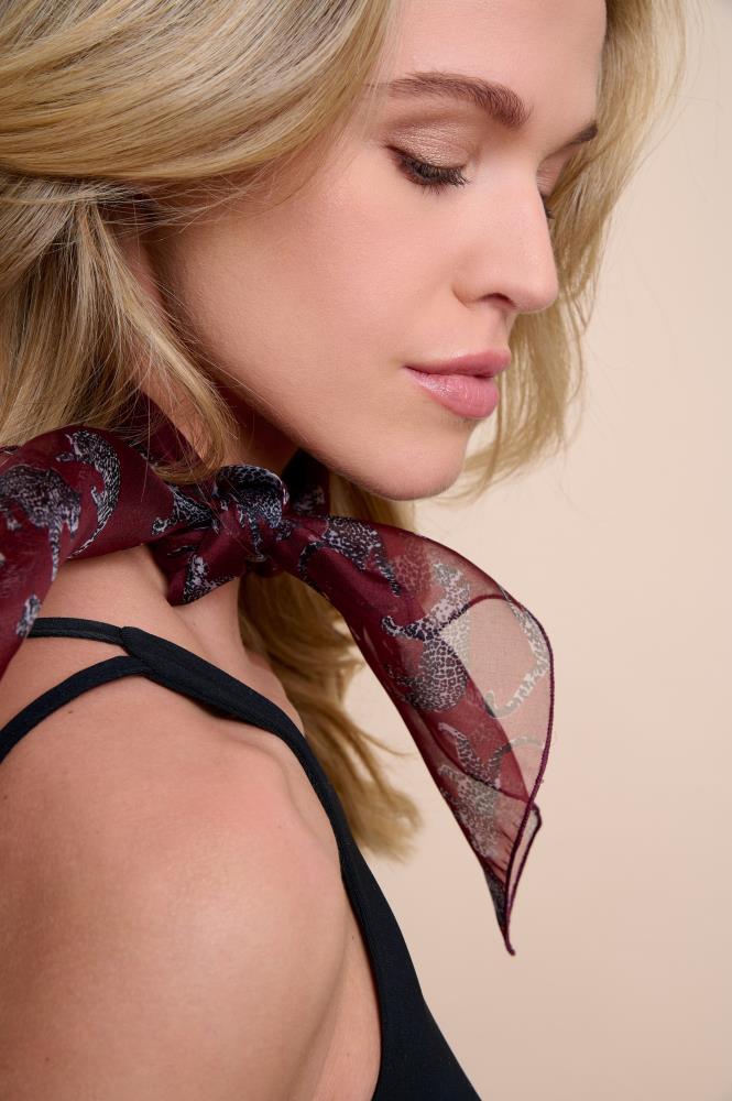 A profile photo of a model focusing on a stylish burgundy silk scarf with a leopard pattern. The scarf is tied around the neck, highlighting the light and sheer fabric texture. Minimalist makeup and soft lighting emphasize the elegant and luxurious image of this accessory.
