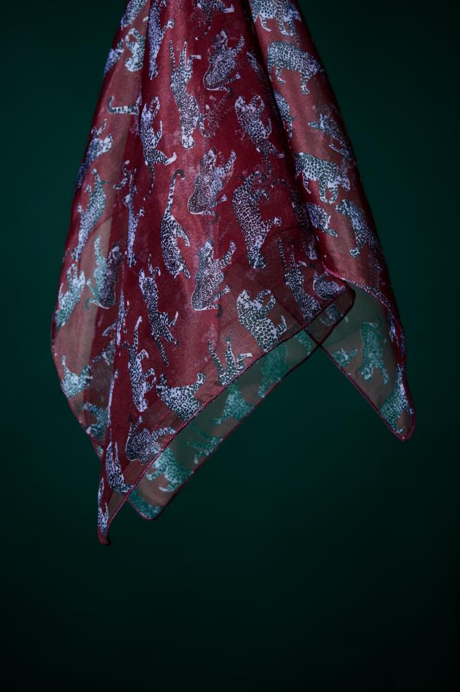 A flowing burgundy silk scarf with a stylish leopard pattern set against a dark green background. The photo highlights the fabric's transparency, subtle sheen, and detailed grey-toned animal motifs.