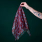 A hand holding a burgundy silk scarf with a stylish leopard pattern against a dark green background. The photo shows a tattooed arm holding the light, sheer fabric, which features a subtle sheen and grey-toned animal motifs.
