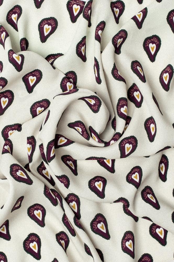 A close-up of crumpled light fabric with a repeating heart pattern. Each heart is yellow, encased in a dark burgundy outline with the words "With LOVE" written above it. The folds swirl in the center, creating a sense of depth and highlighting the softness of the fabric.