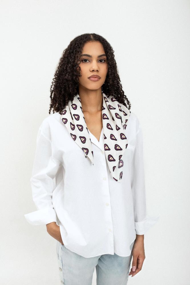 A young woman with dark curly hair poses against a light background, looking directly at the camera. She is wearing a loose-fitting white button-down shirt with slightly rolled sleeves and light blue jeans. A white scarf with a repeating heart pattern is draped over her shoulders, with its ends hanging freely in front. She has one hand in her pocket, and her pose is calm and confident.
