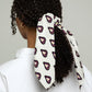 A back view of a young woman with dark curly hair tied in a low ponytail. Her hair is styled with a light-colored scarf featuring a repeating burgundy heart pattern, with its long ends hanging freely. She is wearing a white shirt, and the background is light and neutral.