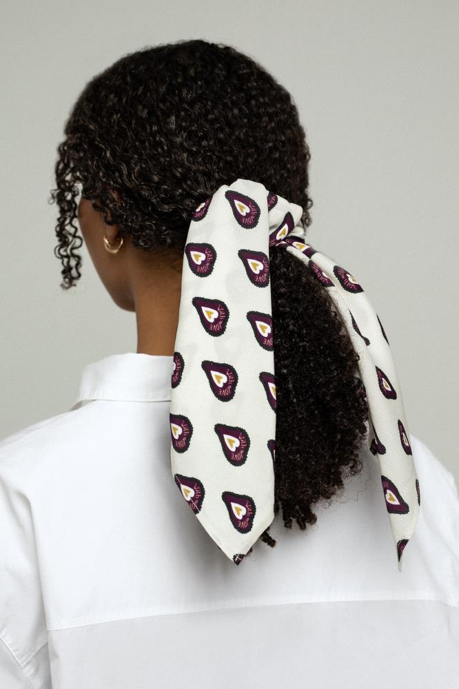 A back view of a young woman with dark curly hair tied in a low ponytail. Her hair is styled with a light-colored scarf featuring a repeating burgundy heart pattern, with its long ends hanging freely. She is wearing a white shirt, and the background is light and neutral.