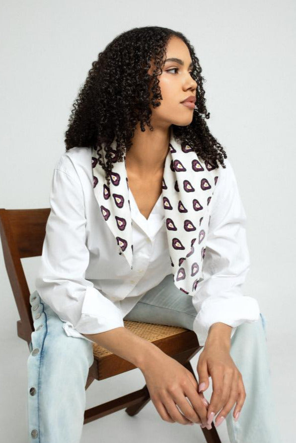 Woman wearing a white shirt with a patterned viscose scarf, sitting on a chair against a plain background