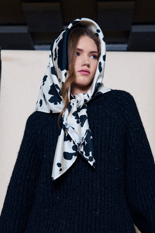 A medium shot of a young woman posing against a studio backdrop, looking to the side. She is wearing a dark, chunky knit sweater, with a silk scarf wrapped around her head and shoulders. The scarf features a bold black and white pattern with irregular spots, reminiscent of a Dalmatian print, and is stylishly tied under her chin. The light background behind the model creates a contrast with the dark outfit, highlighting the graphic nature of the accessory and the modern look.