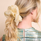 Woman with blonde hair tied back with a large champagne hue silk organza scrunchie against a beige background