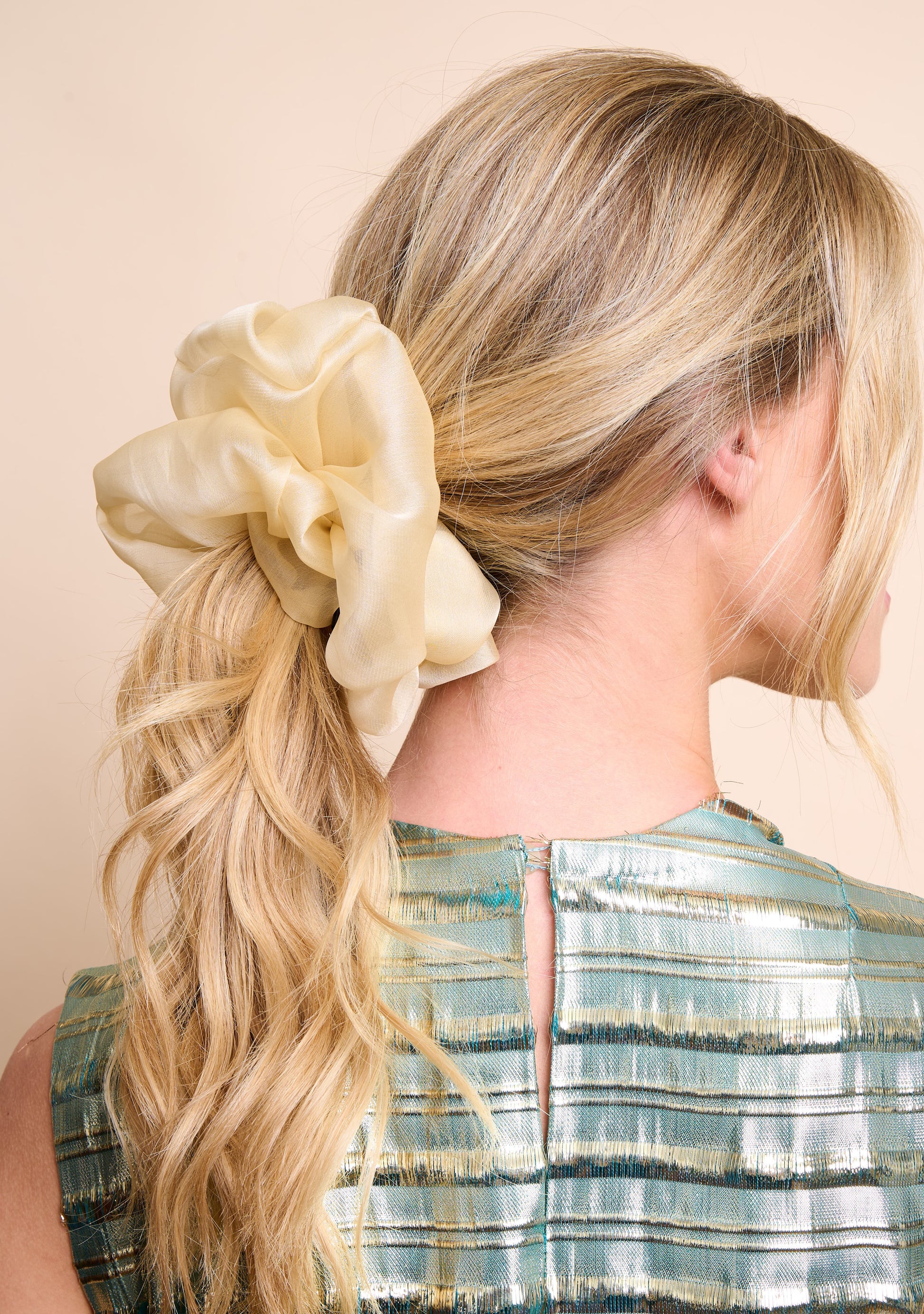 Woman with blonde hair tied back with a large champagne hue silk organza scrunchie against a beige background