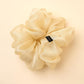 large, voluminous cream-colored or champagne sheer fabric (organza) scrunchie placed against a warm yellowish background. The accessory features abundant, soft folds and a subtle sheen, with a small black brand label visible on the right side.
