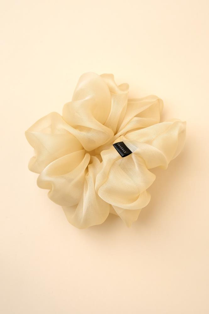 large, voluminous cream-colored or champagne sheer fabric (organza) scrunchie placed against a warm yellowish background. The accessory features abundant, soft folds and a subtle sheen, with a small black brand label visible on the right side.