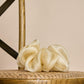 A voluminous, cream-colored or champagne sheer fabric scrunchie placed on the woven seat of a wooden chair. The light fabric folds and subtle sheen give the accessory an elegant, airy look. The warm lighting and natural chair textures create a cozy, rustic atmosphere.
