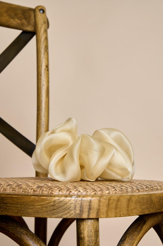 A voluminous, cream-colored or champagne sheer fabric scrunchie placed on the woven seat of a wooden chair. The light fabric folds and subtle sheen give the accessory an elegant, airy look. The warm lighting and natural chair textures create a cozy, rustic atmosphere.