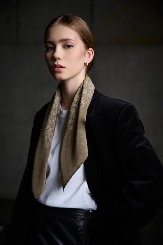 A medium-shot portrait of a woman posing against a dark, industrial-style background. She is wearing a luxurious black velvet blazer, a white t-shirt, and a black leather skirt. A brownish silk scarf, decorated with a subtle light floral pattern, is loosely draped around her neck. Side lighting creates a play of shadows, highlighting the model's features and the contrasting textures of velvet, leather, and silk, giving the look a sophisticated feel.