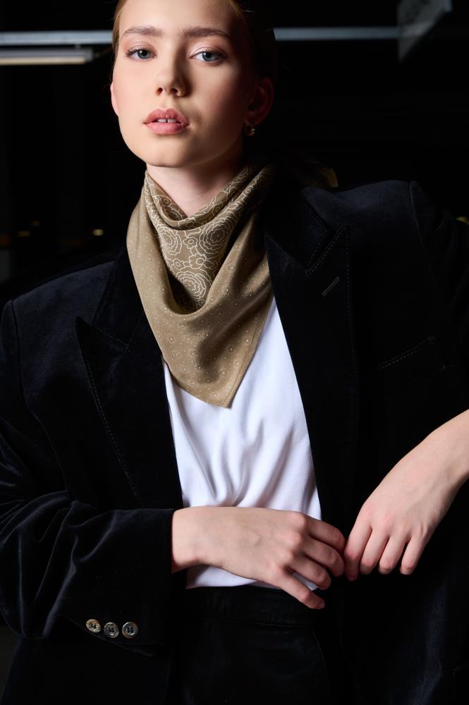a woman posing in a luxurious black velvet blazer and a white t-shirt. A brownish silk scarf, decorated with a fine light pattern, is elegantly tied around her neck. The model looks directly at the camera, with one hand lightly holding the edge of the blazer. The dark background and directional lighting create a classic, sophisticated, and stylish mood, highlighting the velvet texture and the subtle pattern of the accessory.