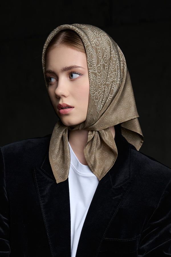 A portrait of a woman posing while looking to the side, wearing a black velvet blazer and a white t-shirt. Her head is covered with a brownish silk scarf decorated with a fine light floral pattern. The scarf is stylishly tied under her chin, with its ends falling freely onto the shoulders of the blazer. The dark background and directional lighting create a classic, sophisticated look, highlighting the texture and elegance of the accessory.