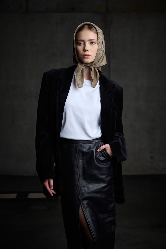 A full-length photo of a woman posing against a concrete wall in an industrial setting. She is wearing a black velvet blazer over a white t-shirt and a black leather skirt with a slit. Her head is covered with a brownish silk scarf, stylishly tied under her chin. The model looks directly at the camera with one hand in her skirt pocket. The composition creates a sophisticated, modern, and slightly edgy look, highlighting the textures of different fabrics – velvet, leather, and silk.