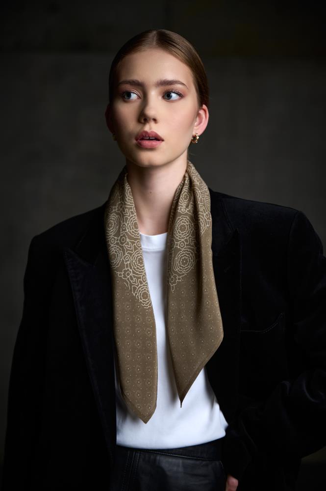 A medium-shot portrait of a woman posing while looking to the side against a dark, neutral background. She is wearing a black velvet blazer, a white t-shirt, and black leather trousers or a skirt. A brownish silk scarf, decorated with a fine light floral pattern, is loosely draped around her neck. Soft, directional lighting highlights the model's features and the luxurious texture of velvet and silk, creating a stylish and sophisticated look.