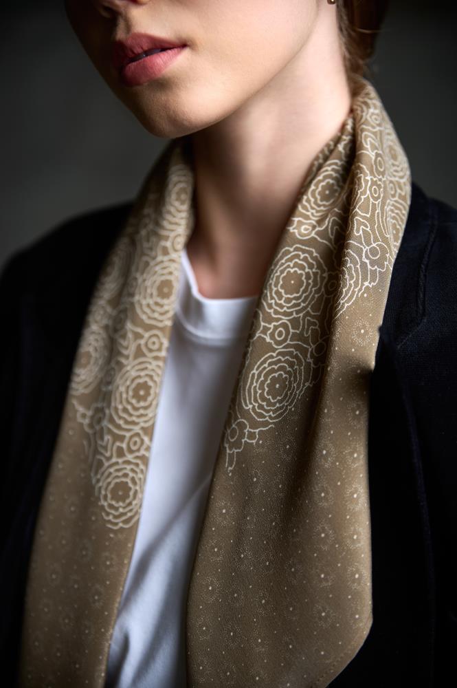 A close-up photo focusing on a brownish silk scarf adorning a woman's neck and shoulders. The scarf is decorated with a fine, white floral and dot pattern and is loosely draped over a white t-shirt and a black blazer. Soft, directional lighting highlights the texture of the fabric and the subtle detail of the pattern, creating a calm and elegant aesthetic.