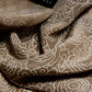 An extreme close-up (macro) showing the folds of a brownish silk scarf. The fabric is decorated with a fine light floral and dot pattern. A black label with the text "SKARELE" is visible in the upper part of the photo. Soft lighting highlights the subtle sheen of the fabric, the smooth material texture, and how the fabric naturally drapes and plays with light, creating a sense of luxury and softness.