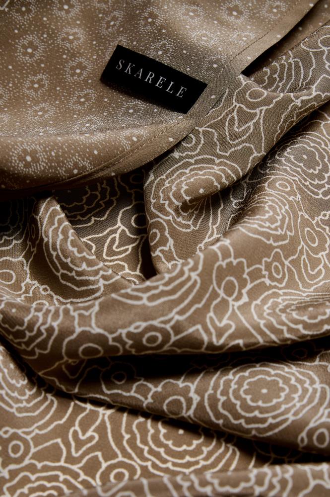 An extreme close-up (macro) showing the folds of a brownish silk scarf. The fabric is decorated with a fine light floral and dot pattern. A black label with the text "SKARELE" is visible in the upper part of the photo. Soft lighting highlights the subtle sheen of the fabric, the smooth material texture, and how the fabric naturally drapes and plays with light, creating a sense of luxury and softness.