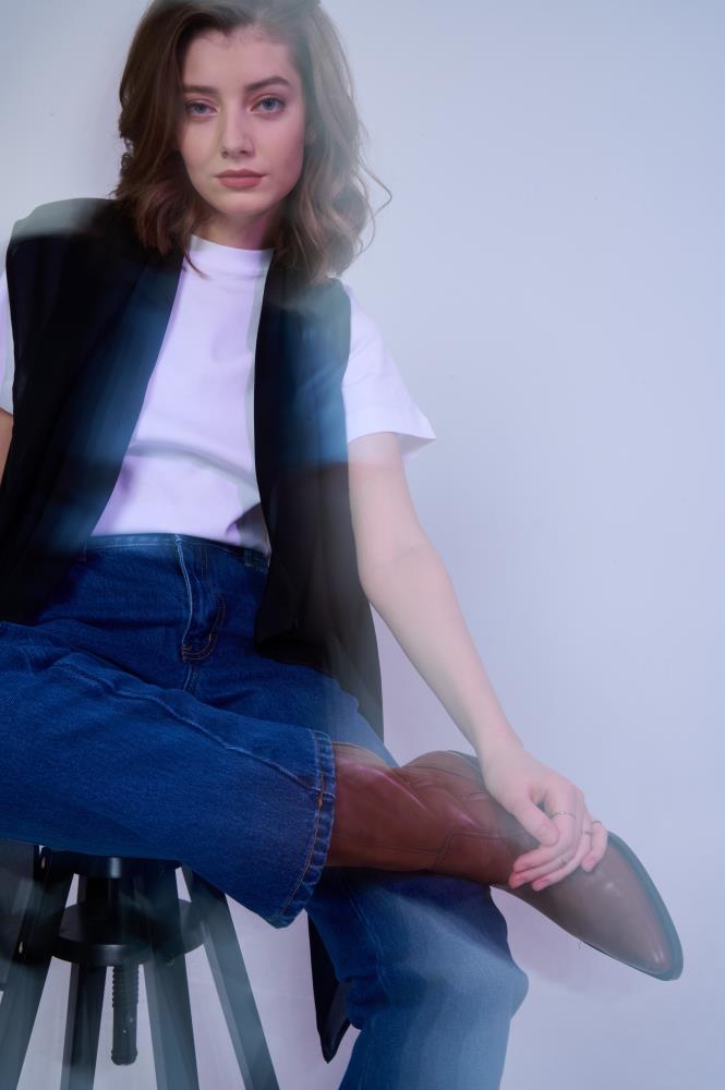 A long, sheer black scarf layered over a white t-shirt and blue jeans. The model is seated, highlighting the garment's length and transparency. Soft, high-key lighting emphasizes the casual yet stylish outfit.