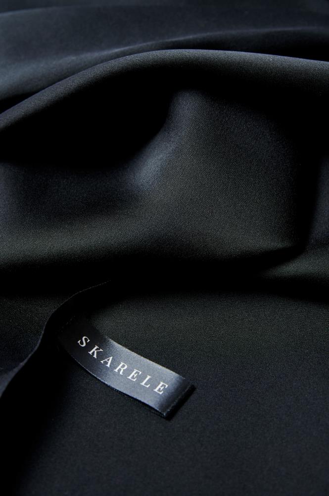 A macro photograph of a black silk scarf showing deep, soft folds and a subtle matte sheen. A small black satin label with the brand name "SKARELE" in white capital letters is stitched onto the fabric, highlighting the premium quality and texture of the material.